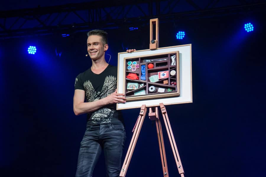 magier-auftritt-zaubershow Zauberer hält Bild mit verschiedenen Objekten auf Bühne.